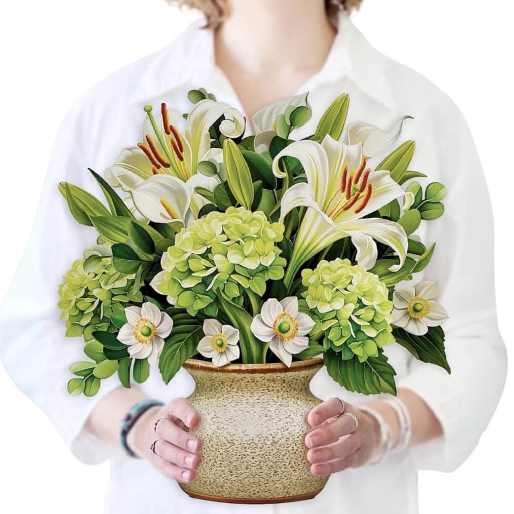 A person in a white shirt holds a decorative vase with a bouquet of white lilies and green hydrangeas, evoking the elegance of easy flower paper crafts. The focus is on the vibrant flowers against a plain background.